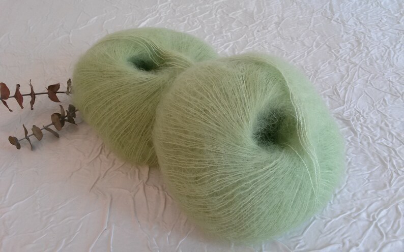 May include: Two skeins of light green yarn, likely mohair or a similar soft fiber, are shown on a white textured surface. The yarn is wound into balls and ready for knitting or crocheting.