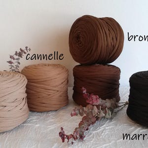 May include: An assortment of five large, round balls of fabric yarn in various shades of brown and beige. The colors are labeled "beige", "cannelie", "bronze", and "marron fonce". The yarn is arranged on a white surface with dried floral accents.