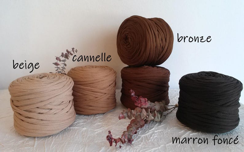 May include: An assortment of five large, round balls of fabric yarn in various shades of brown and beige. The colors are labeled "beige", "cannelie", "bronze", and "marron fonce". The yarn is arranged on a white surface with dried floral accents.