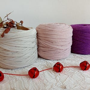 May include: Three spools of yarn in different colours: beige, pink, and purple. The yarn is made of a soft, stretchy material and has a subtle shimmer.