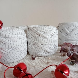 May include: Three white yarn balls with a woven pattern. The yarn is white with a thin black stripe. The yarn balls are on a white surface with red bells and a brown leaf.