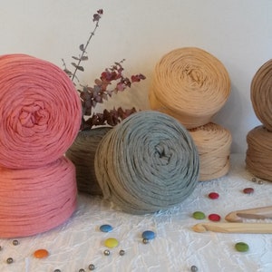 Op de afbeelding: Vijf spoelen garen in verschillende kleuren, waaronder roze, beige, bruin en grijs. Het garen is in een cirkel op een wit oppervlak gerangschikt met kleurrijke snoepjes en zilveren kralen die eromheen liggen.