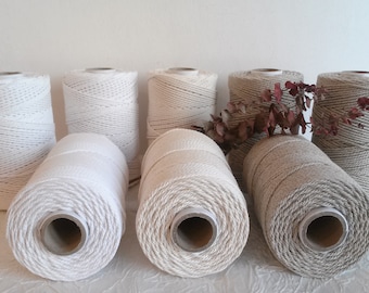 May include: Spools of macrame cord in various neutral colors, including white, cream, and gray. The spools are cylindrical and made of tightly wound cotton cord. A few dried floral stems are placed between the spools.