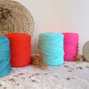 May include: Five large spools of colorful t-shirt yarn in teal, red, turquoise, pink, and light green. The yarn is arranged on a white surface with decorative elements in the background, suitable for crafting projects.