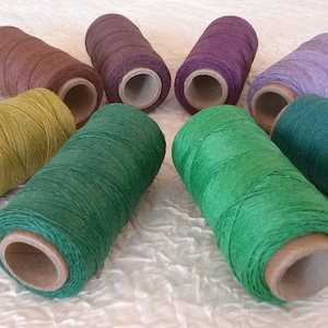 May include: Eight spools of waxed linen thread in various colours, including brown, green, yellow, purple, and blue. The spools are arranged in a circle on a white textured surface.