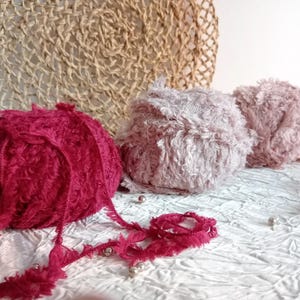 Puede incluir: Tres ovillos de lana en diferentes colores: rojo, rosa y un tono rosa más claro. La lana es esponjosa y tiene un aspecto texturizado.