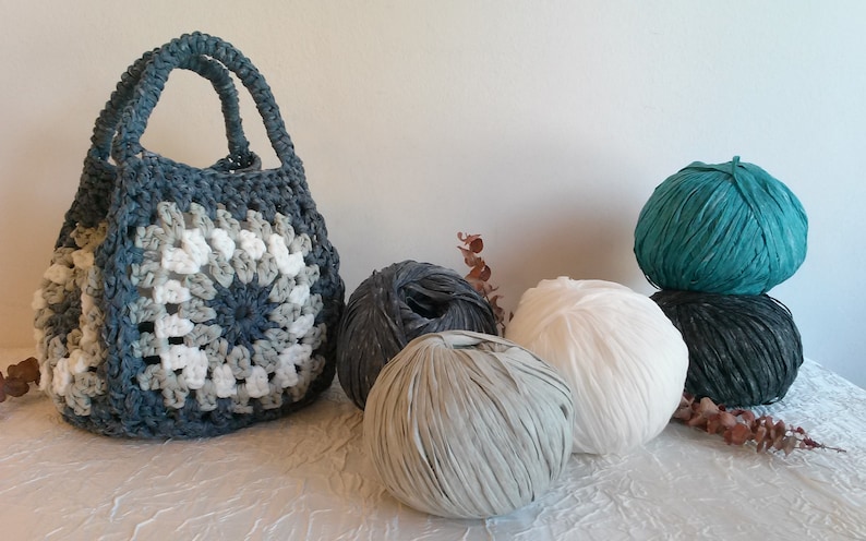 May include: A crocheted tote bag with a blue, white, and gray pattern. Four balls of yarn in various colors are displayed next to the bag.