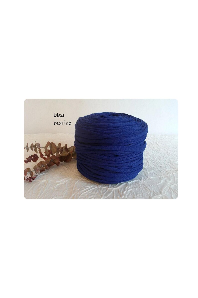 May include: A tightly wound roll of navy blue fabric, possibly jersey knit, sits on a white surface. The text "bleu marine" is visible in the upper left corner. Dried foliage adds a natural accent.