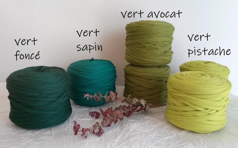 May include: A collection of green fabric yarn in various shades, including dark green, teal, and avocado green. The yarn is arranged in neat, round bundles, with labels indicating the color names. A sprig of dried leaves adds a natural touch.
