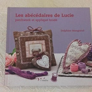 May include: A book titled "Les abécédaires de Lucie" with a purple and brown patchwork heart and a pillow with a similar design. The book cover also features embroidery and the text "patchwork et appliqué brodé" and "Delphine Muggrand."