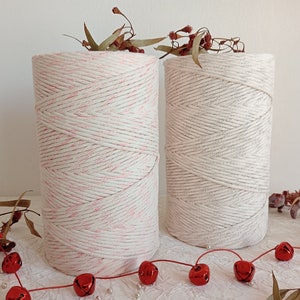 Puede incluir: Dos carretes de cordón de algodón natural, uno blanco y otro con un patrón de rayas rosas. Los carretes están sobre una superficie blanca con cascabeles rojos esparcidos alrededor.