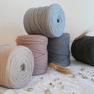 Puede incluir: Cinco carretes de hilo grueso y esponjoso en varios colores, incluyendo gris claro, rosa, gris oscuro y verde oscuro. Los carretes están dispuestos en forma de pirámide sobre una superficie blanca.