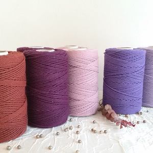 Puede incluir: Cinco carretes de cuerda de algodón en varios tonos de morado, rosa y marrón. Los carretes están dispuestos en una fila sobre una superficie blanca.