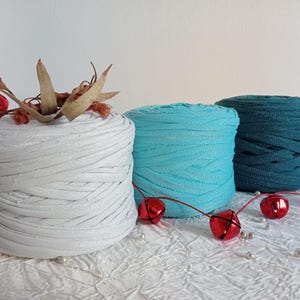 May include: Three spools of yarn in white, turquoise, and teal. The yarn is thick and textured, and the spools are arranged in a row on a white surface. There are red bells and sprigs of dried eucalyptus scattered around the spools.