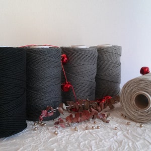 May include: Five spools of cotton rope in black, gray, and natural colors. The spools are arranged in a row on a white textured surface. Red bells and a sprig of eucalyptus are scattered around the spools.