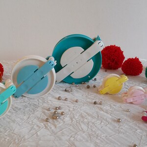 May include: Assortment of colourful pom-pom makers in various sizes. The makers are made of white and blue plastic with a handle. There are also several colourful pom-poms made of yarn.