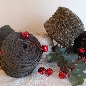 Puede incluir: Una variedad de ovillos de hilo en tonos grises, negros y beige, con una guirnalda de campanas rojas y ramitas de vegetación. El hilo tiene un brillo metálico, y la escena está ambientada sobre un fondo blanco, creando una exhibición festiva.