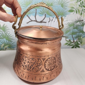 May include: A copper-colored pot with a lid and a decorative handle. The pot features etched floral and geometric patterns around the body. The handle and the lid's knob are gold-colored.