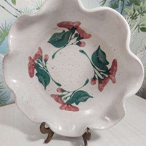 May include: A white ceramic serving bowl with a scalloped edge, speckled surface, and floral design. The bowl features red flowers and green leaves arranged in a circular pattern. It is displayed on a bronze-colored metal stand.