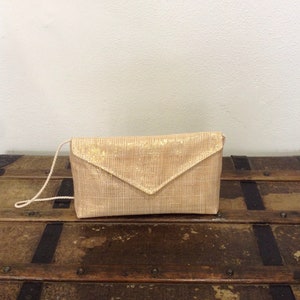 Op de afbeelding: Een lichtgouden clutch met een geweven textuur en een driehoekige klepsluiting. De clutch heeft een dunne, bijpassende schouderriem. De tas ligt op een houten oppervlak.