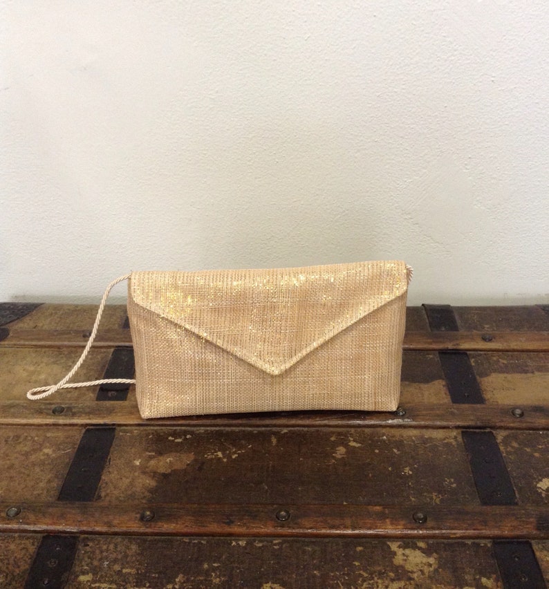 Op de afbeelding: Een lichtgouden clutch met een geweven textuur en een driehoekige klepsluiting. De clutch heeft een dunne, bijpassende schouderriem. De tas ligt op een houten oppervlak.