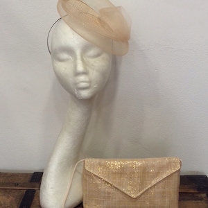 Op de afbeelding: Een perzikkleurige hoed en bijpassende clutch. De hoed, op een mannequinhoofd, heeft een transparant, gelaagd ontwerp. De clutch heeft een getextureerde, glinsterende afwerking en een dunne riem. De items worden gepresenteerd op een houten oppervlak.