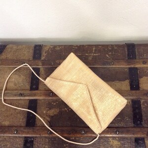 Op de afbeelding: Een beige envelop clutch met een geweven textuur en een bijpassende touwriem. De tas is rechthoekig met een driehoekige klepsluiting. De clutch ligt op een vintage houten koffer.