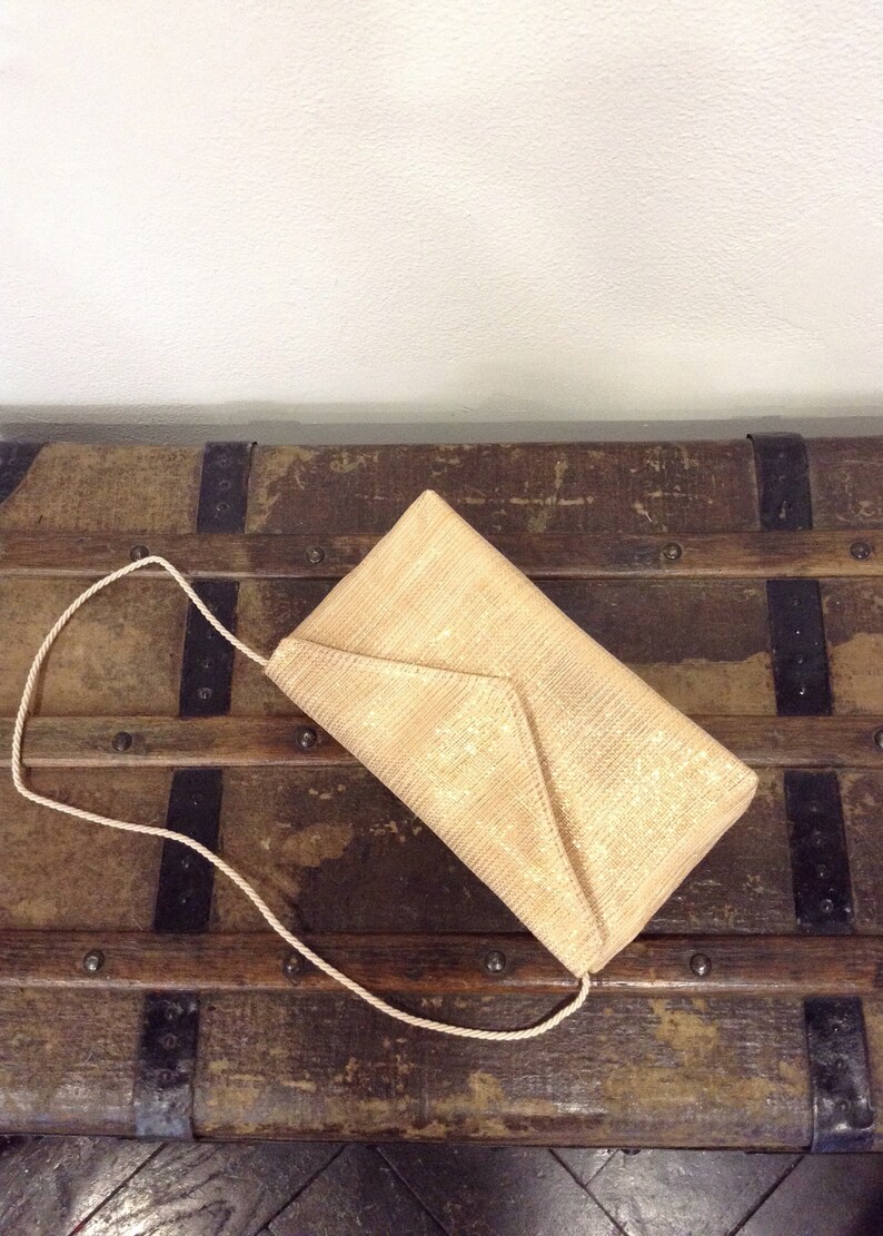 Op de afbeelding: Een beige envelop clutch met een geweven textuur en een bijpassende touwriem. De tas is rechthoekig met een driehoekige klepsluiting. De clutch ligt op een vintage houten koffer.