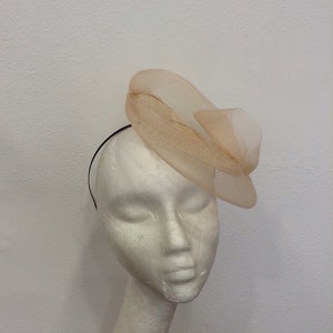 Op de afbeelding: Een perzikkleurige fascinator met een gelaagd, doorschijnend ontwerp en een zwarte haarband. Het hoofddeksel heeft een gestructureerde, gebogen vorm met een delicate, luchtige uitstraling. De fascinator is bevestigd aan een zwarte haarband.
