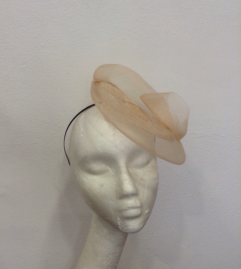 Op de afbeelding: Een perzikkleurige fascinator met een gelaagd, doorschijnend ontwerp en een zwarte haarband. Het hoofddeksel heeft een gestructureerde, gebogen vorm met een delicate, luchtige uitstraling. De fascinator is bevestigd aan een zwarte haarband.