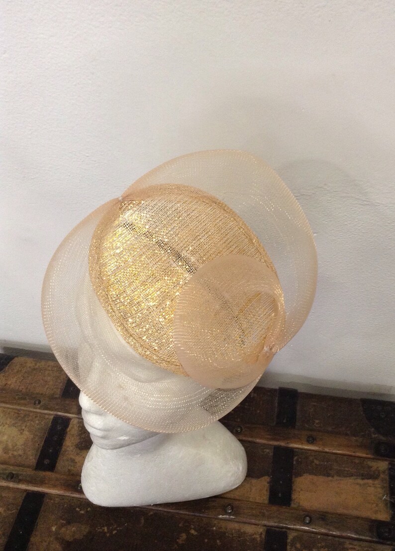 Op de afbeelding: Perzik kleurige formele hoed met een gelaagd ontwerp. De hoed heeft een gouden, getextureerd midden en wordt getoond op een witte mannequin hoofd. Het ontwerp van de hoed omvat een brede rand en een decoratieve, wervelende bovenkant.