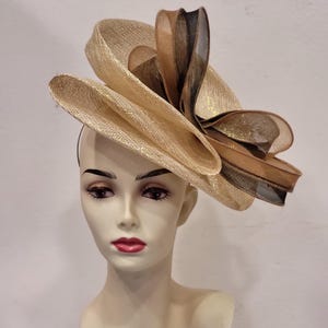 Peut inclure: Un chapeau décoratif doré avec un grand design en couches. Le chapeau présente un large bord et est orné d'accents marron et noirs. Le chapeau est présenté sur une tête de mannequin.