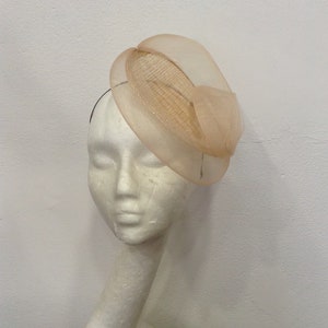 Op de afbeelding: Een beige fascinator hoed met een cirkelvormige basis en een doorschijnende, gedrapeerde overlay. De hoed wordt getoond op een witte mannequinhoofd, wat het elegante ontwerp laat zien. Perfect voor bruiloften of speciale evenementen.