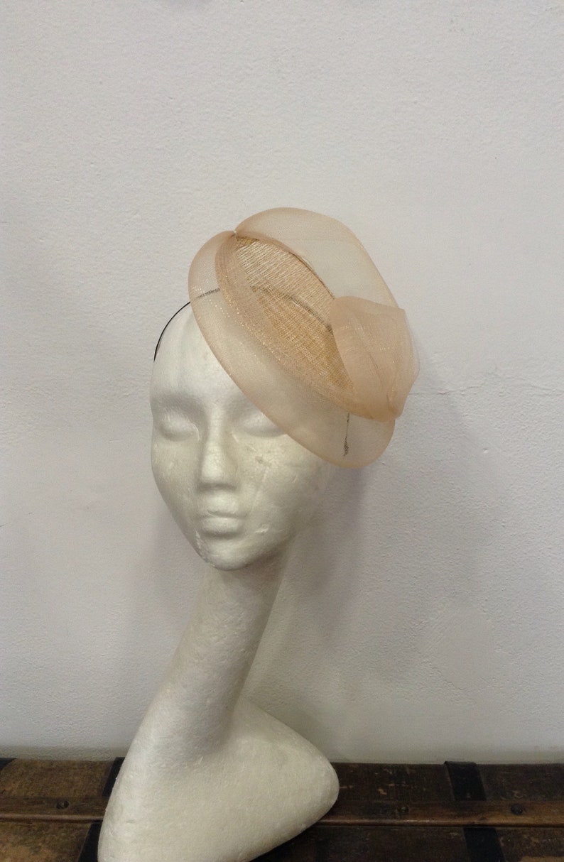 Op de afbeelding: Een beige fascinator hoed met een cirkelvormige basis en een doorschijnende, gedrapeerde overlay. De hoed wordt getoond op een witte mannequinhoofd, wat het elegante ontwerp laat zien. Perfect voor bruiloften of speciale evenementen.