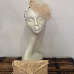 Op de afbeelding: Een perzikkleurige hoed met een gelaagd, doorschijnend ontwerp staat op een mannequinhoofd. Een bijpassende clutch met een gouden glitterafwerking ligt eronder. De items staan op een houten kist, wat wijst op een vintage of formele thema.