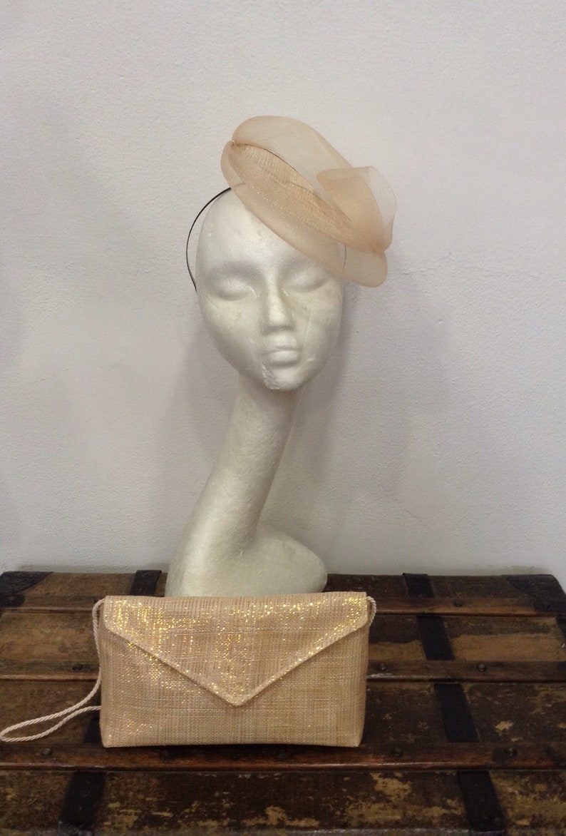 Op de afbeelding: Een perzikkleurige hoed met een gelaagd, doorschijnend ontwerp staat op een mannequinhoofd. Een bijpassende clutch met een gouden glitterafwerking ligt eronder. De items staan op een houten kist, wat wijst op een vintage of formele thema.