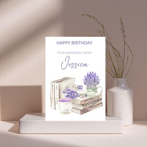 May include: A birthday card featuring a watercolour illustration of books, lavender, and a teacup. The card reads "Happy Birthday To My Wonderful Sister Jessica" in purple script. The card is on a white plinth.