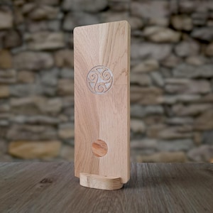 Könnte beinhalten: Ein Handy-Ständer aus Holz mit einem keltischen Knoten-Design, das oben eingraviert ist. Der Ständer ist aus hellfarbenem Holz gefertigt und hat einen kleinen, abgerundeten Sockel. Der Hintergrund ist eine Steinmauer.