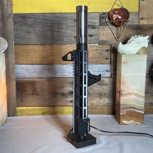 May include: A black and silver lamp with a unique design, resembling a rifle barrel. The lamp has a rectangular base and a cylindrical top, with a series of rectangular cutouts along the sides. A black handle is attached to the side.