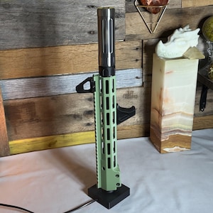 May include: A green and black lamp with a cylindrical top and a rectangular base. The lamp has a series of vertical slots along its sides and a handle-like feature. The lamp is plugged into an electrical outlet.