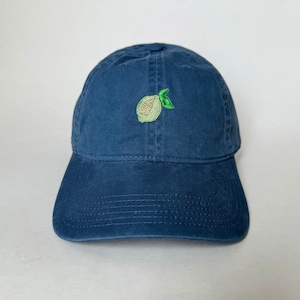 Puede incluir: Gorra de béisbol azul marino con visera curva. La gorra presenta un diseño bordado de una fruta verde claro y amarilla en la parte delantera. El sombrero está hecho de una tela suave y texturizada.
