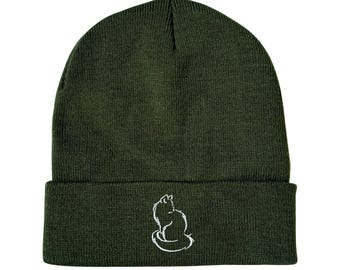 Puede incluir: Un gorro de lana verde oscuro con un contorno blanco de un gato sentado en la parte delantera.