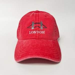 Puede incluir: Gorra de béisbol roja con visera curva. La gorra presenta un diseño bordado del Tower Bridge en blanco y negro, con la palabra "LONDON" bordada debajo en blanco. La gorra está hecha de una tela suave y lavada.