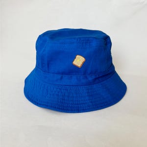May include: A vibrant blue bucket hat featuring an embroidered toast and butter design. The hat is made from a soft material and has a traditional bucket hat silhouette. The hat is a solid blue colour.