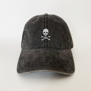 May include: A dark grey, distressed baseball cap featuring a white embroidered skull and crossbones emblem. The cap has a curved peak and a fabric strap. The hat is a dark grey colour.