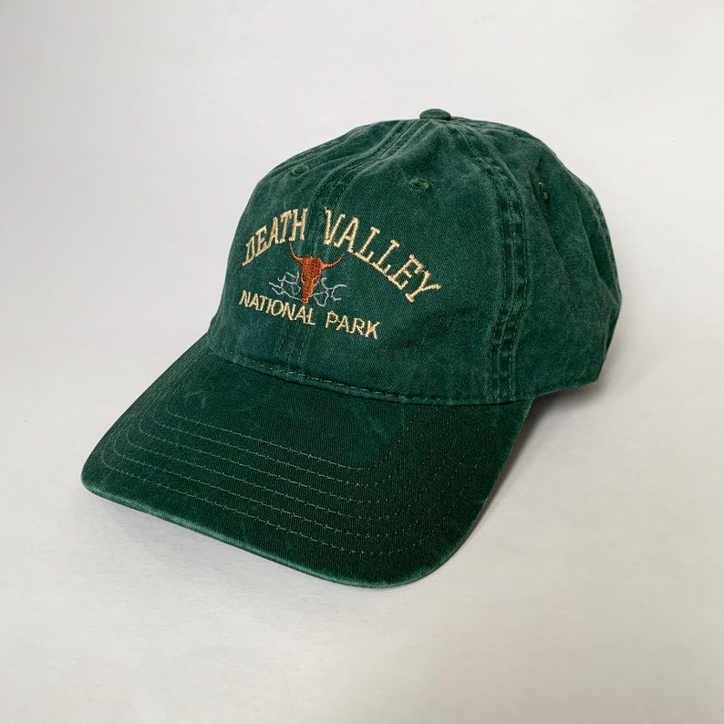 May include: A dark green baseball cap with the text "Death Valley National Park" embroidered on the front. The text is in a white font with a brown bull skull graphic.
