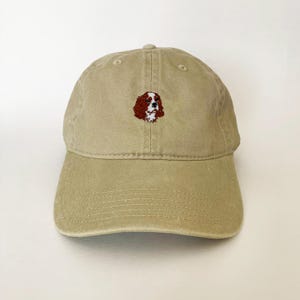 Könnte beinhalten: Beige Baseball Cap mit gebogenem Schirm. Die Kappe hat ein gesticktes Bild eines Cavalier King Charles Spaniel Hundes in Braun, Weiß und Schwarz. Die Kappe ist aus einem weichen, strapazierfähigen Stoff gefertigt.