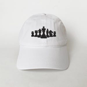 Puede incluir: Gorra de béisbol blanca con un diseño bordado en negro de piezas de ajedrez. El diseño incluye un rey, una reina, una torre, un alfil, un caballo y un peón. La gorra tiene una visera curva y una correa ajustable.