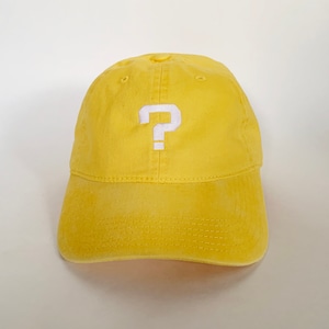 Könnte beinhalten: Gelbe Baseballkappe mit einem weißen, gestickten Fragezeichen auf der Vorderseite. Die Kappe hat einen gebogenen Schirm und ein klassisches Sechs-Panel-Design. Der Stoff scheint eine weiche Baumwollmischung zu sein.