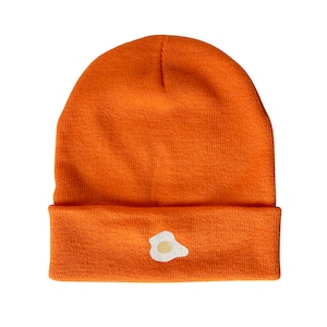 May include: An orange beanie with a white embroidered fried egg design.